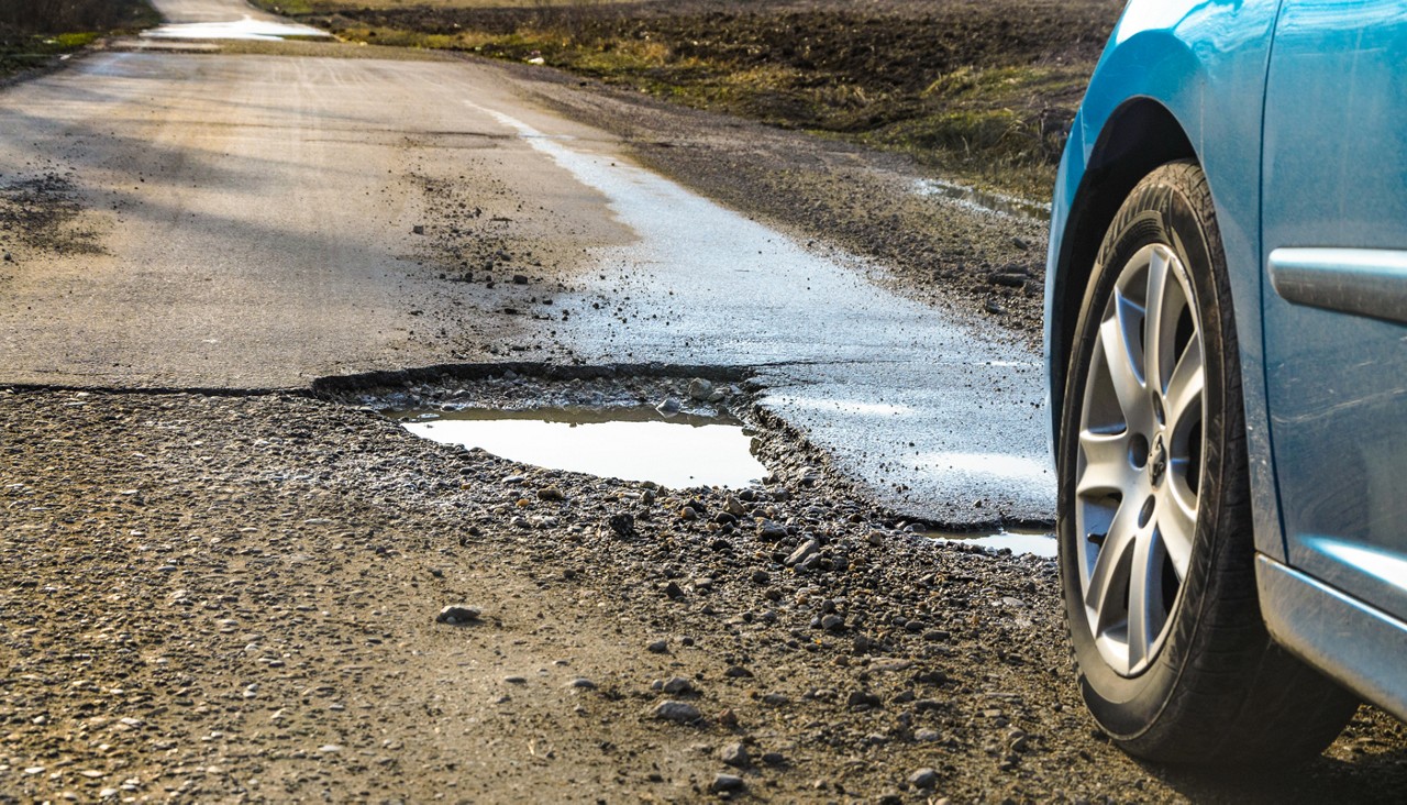 Close up view of a blue car's front tire about to run over a large open pothole.