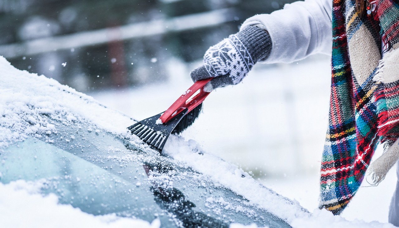 A person scrapping the snow and ice off the windshield of a car in the snow.