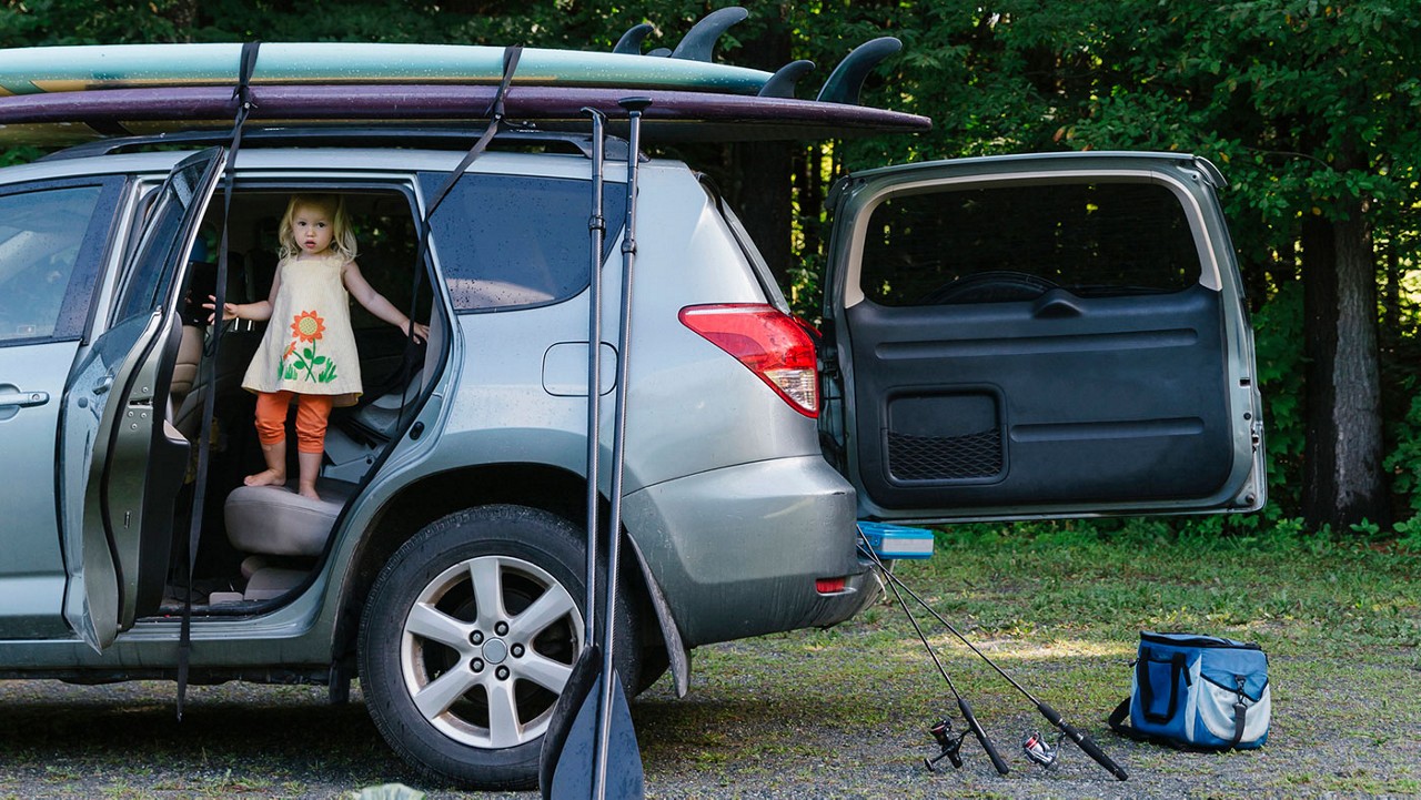 A little girl standing in an open car door as her parents remove camping, fishing and other equipment from the car.