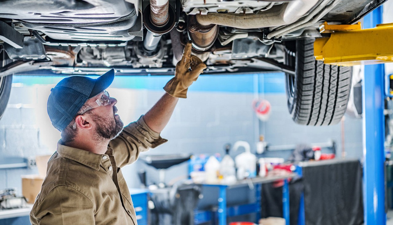 A mechanic with a baseball hat on checking the catalytic converter on a car from underneath.