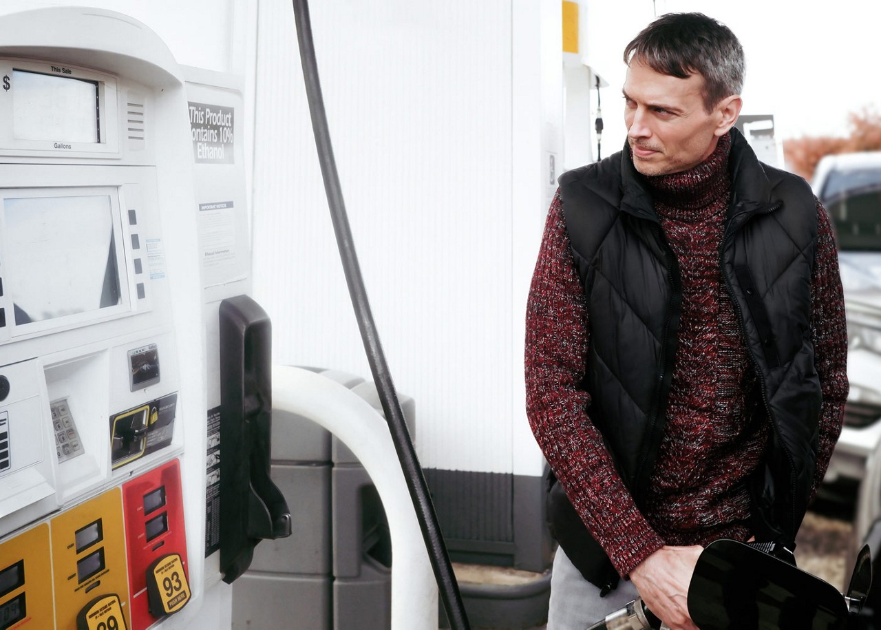 Picture of a man wearing a sweater and vest filling up a gas tank of car.