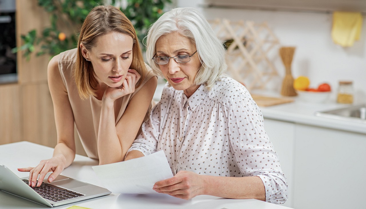 A younger woman on a computer helping an older woman with paperwork.
