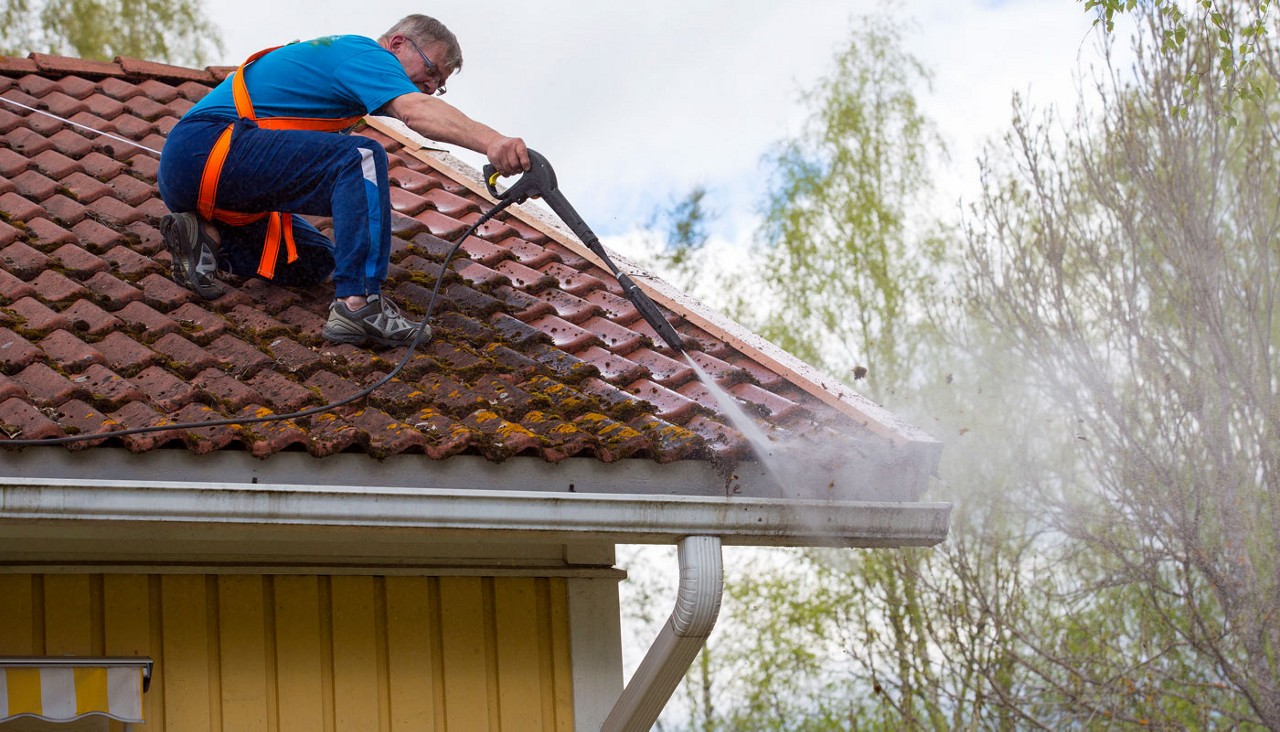 Caucasian man is washing the roof with a high pressure washer. He is wearing safety harness on a slippery roof.