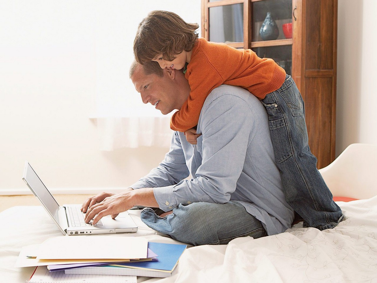A father typing on a laptop while his child hugs him from behind and looks over his shoulder.