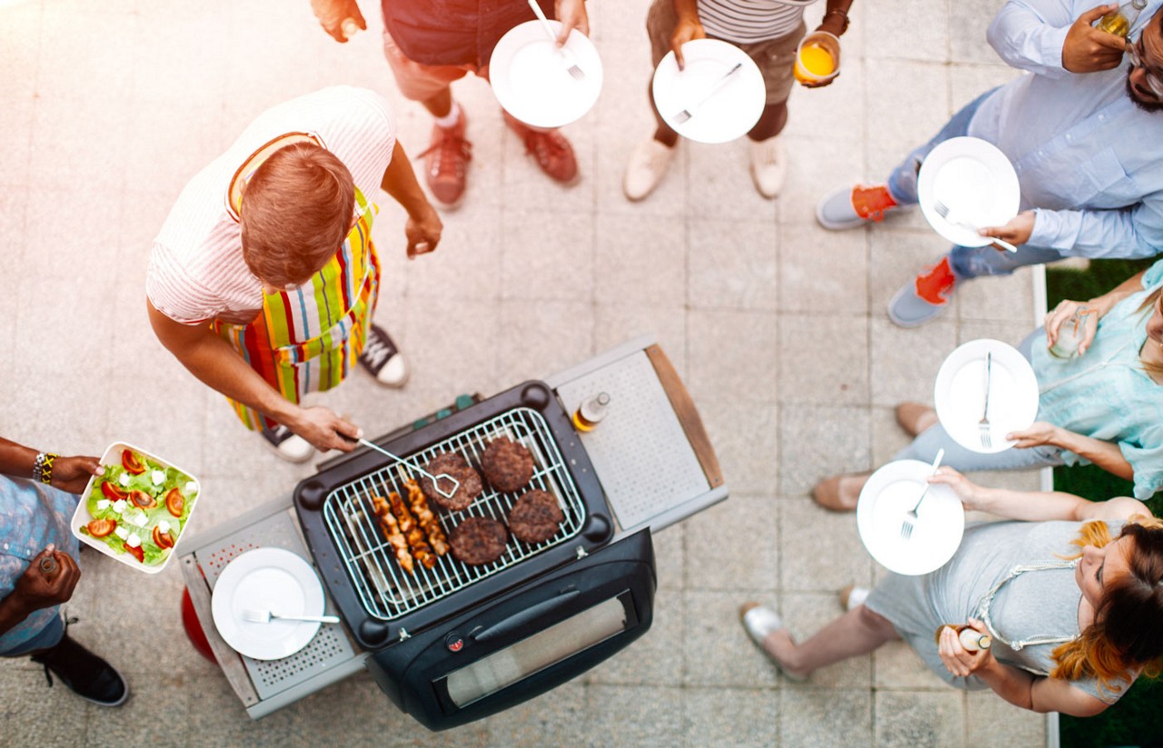 Looking down on a man grilling meat with his friends and family standing around with plates ready to eat.