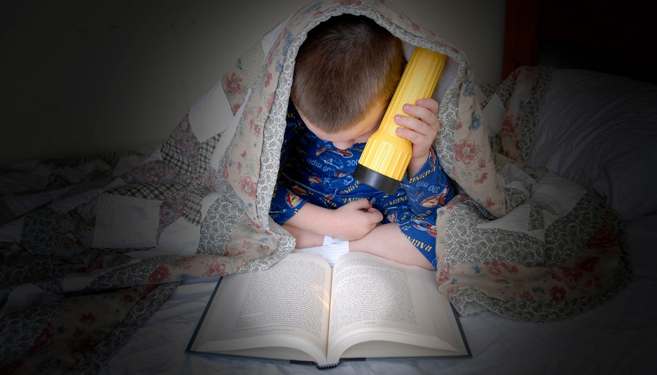 A young boy reading a book with a flashlight under his bed covers