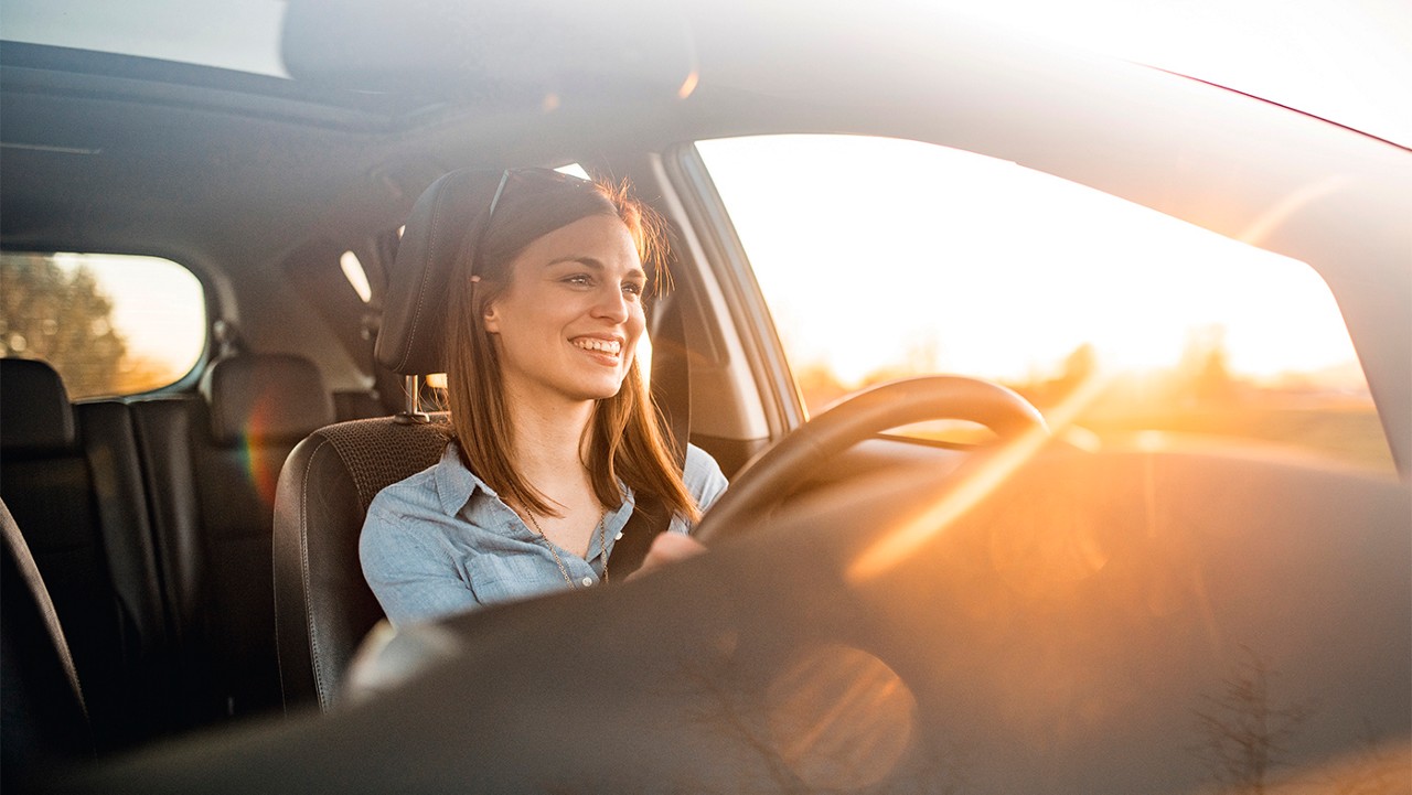 Young woman in a car. She is driving, smiling and looking out. Sun is shining from back on a sunny spring afternoon.