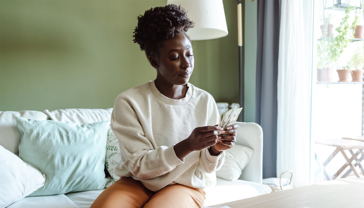 A woman sits on her couch and counts the cash in her hands. 