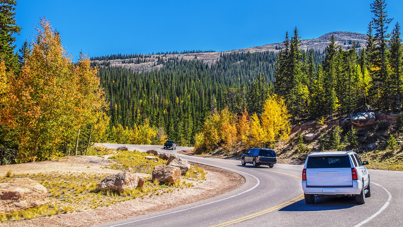 SUVs and pickup truck take sharp turns down mountainside with evergreens and Autumn aspens and bare peaks in background on sunny day