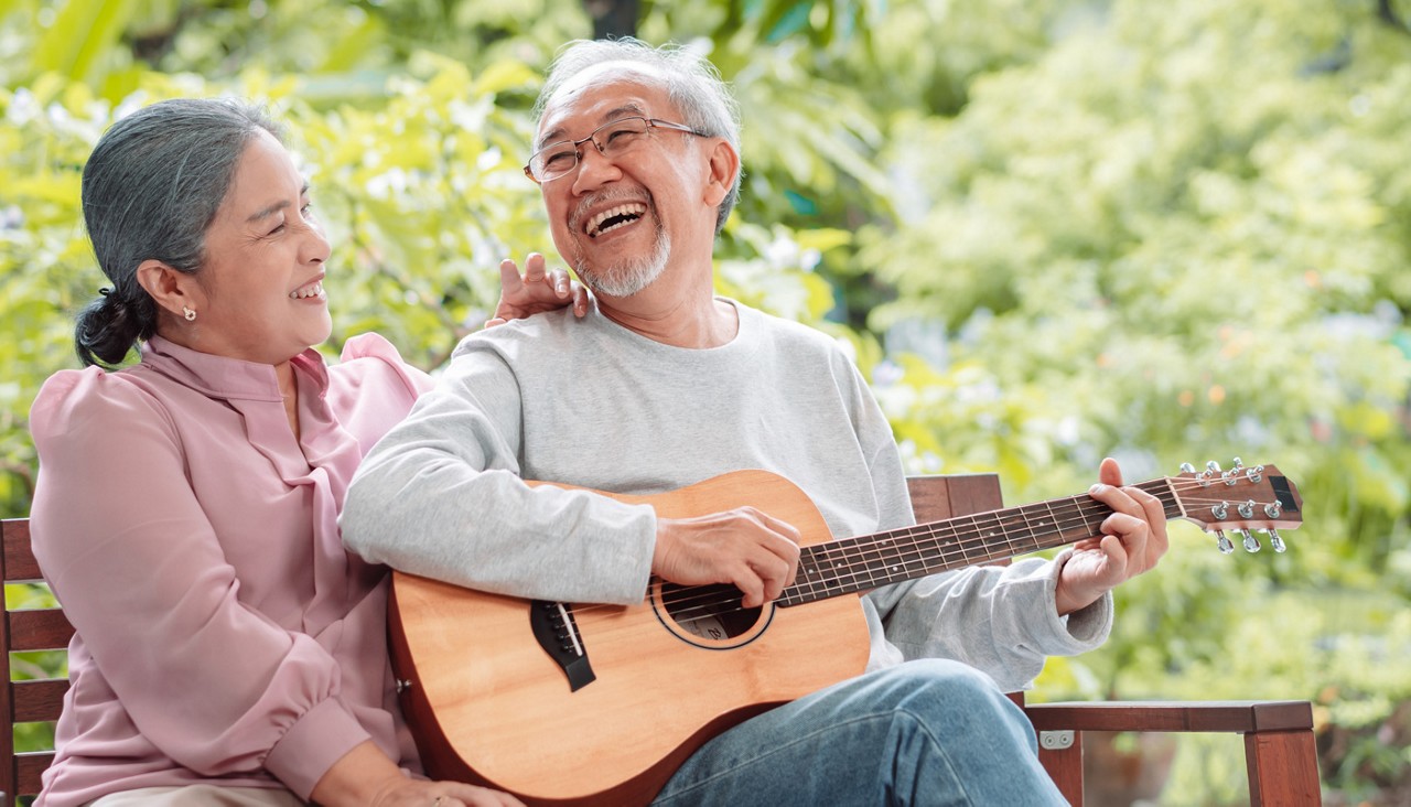 Happy Asian senior couple on park bench, man playing guitar, woman smiling with hand on his shoulder