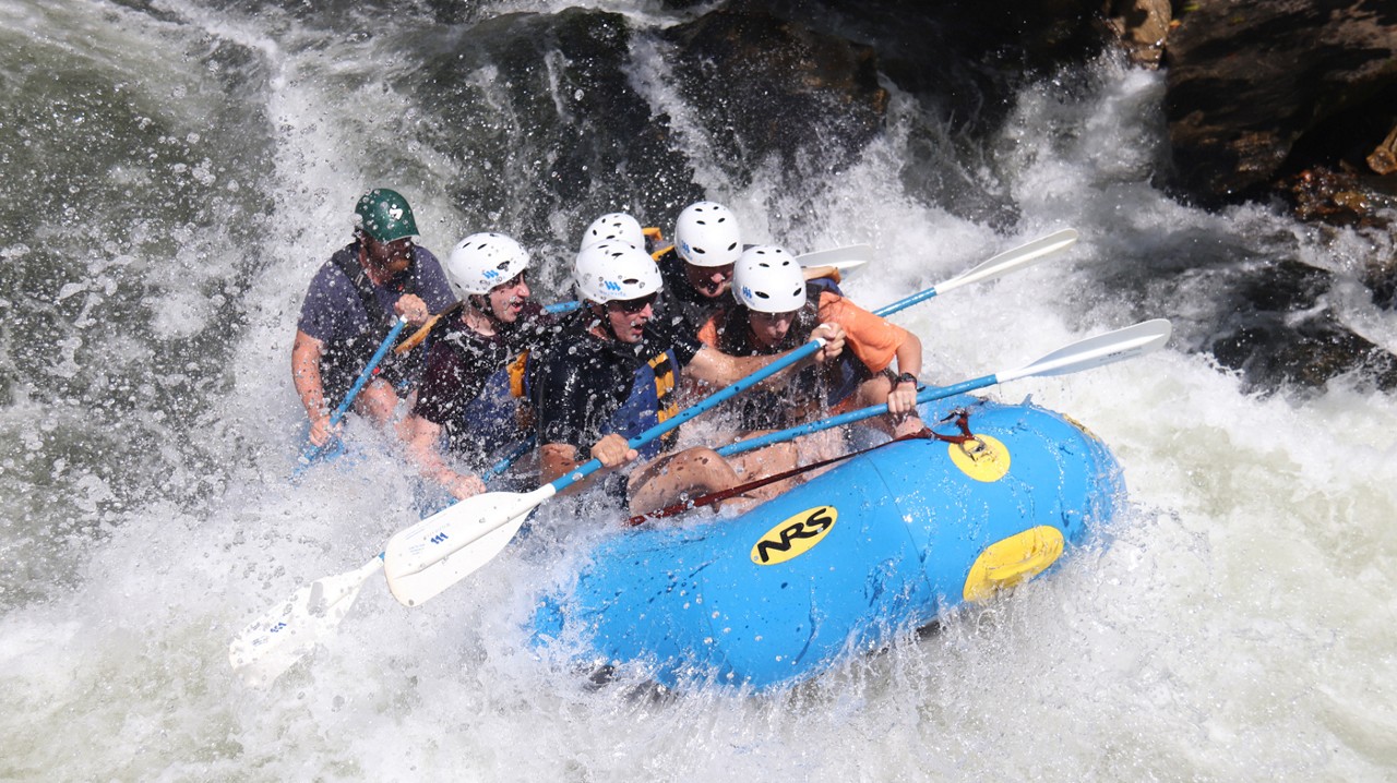 A group of people in safety helmets, t-shirts and shorts in a blue raft going through some rapids on the National Wild & Scenic Chattooga River.